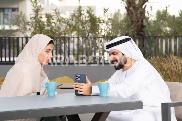 الاستمتاع بشرب المشروبات الساخنه ، رجل عربي يقوم بإلتقاط صورة سيلفي تذكارية له و لزوجته ،  ايماءات السرور والسعادة ، مفهوم المودة والالفة ، قضاء أوقات ممتعة، زوجان عربيان خليجيان اماراتيان مبتسمان يجلسان في المقهى ، قضاء شهر العسل