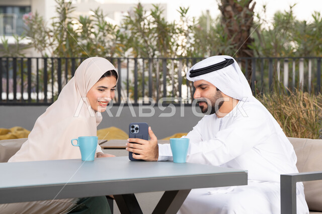 ايماءات السرور والسعادة ، قضاء شهر العسل، الاستمتاع بشرب المشروبات الساخنه ، رجل عربي يقوم بإلتقاط صورة سيلفي تذكارية له و لزوجته ، مفهوم المودة والالفة ، قضاء أوقات ممتعة، زوجان عربيان خليجيان اماراتيان مبتسمان يجلسان في المقهى