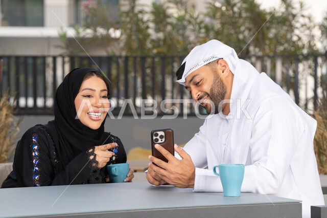 ايماءات السرور والسعادة ، مفهوم المودة والالفة ، قضاء أوقات ممتعة، زوجان عربيان خليجيان اماراتيان مبتسمان يجلسان في المقهى ، يستمتعان بشرب القهوة الساخنة ، رجل عربي يقوم بإلتقاط صورة سيلفي تذكارية له و لزوجته ، الاستمتاع بالاجواء العائلية ، قضاء شهر العسل