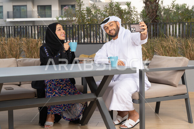 الاستمتاع بالاجواء العائلية ، قضاء شهر العسل، مفهوم المودة والالفة ، قضاء أوقات ممتعة، زوجان عربيان خليجيان اماراتيان مبتسمان يجلسان في المقهى ، يستمتعان بشرب القهوة الساخنة ، رجل عربي يقوم بإلتقاط صورة سيلفي تذكارية له و لزوجته بسعادة