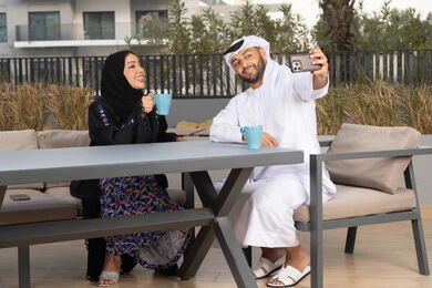 Enjoying the family atmosphere, spending the honeymoon, the concept of affection and intimacy, spending a good time, a smiling Emirati Gulf Arab couple sitting in a cafe, enjoying drinking hot coffee, an Arab man happily taking a souvenir selfie of him and his wife