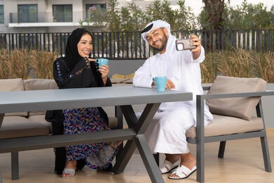قضاء شهر العسل، مفهوم المودة والالفة ، قضاء أوقات ممتعة، زوجان عربيان خليجيان اماراتيان مبتسمان يجلسان في المقهى ، يستمتعان بشرب القهوة الساخنة ، رجل عربي يقوم بإلتقاط صورة سيلفي تذكارية له و لزوجته بسعادة
