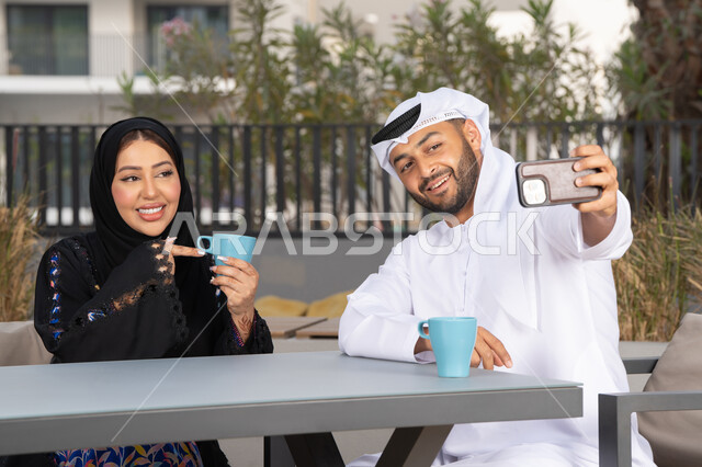 مفهوم المودة والالفة ، قضاء أوقات ممتعة، زوجان عربيان خليجيان اماراتيان مبتسمان يجلسان في المقهى ، يستمتعان بشرب القهوة الساخنة ، رجل عربي يقوم بإلتقاط صورة سيلفي تذكارية له و لزوجته بسعادة ، قضاء شهر العسل