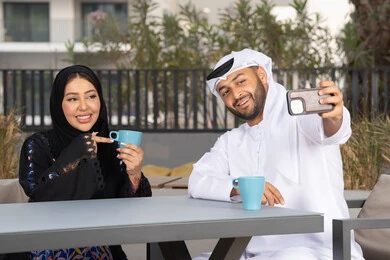 مفهوم المودة والالفة ، قضاء أوقات ممتعة، زوجان عربيان خليجيان اماراتيان مبتسمان يجلسان في المقهى ، يستمتعان بشرب القهوة الساخنة ، رجل عربي يقوم بإلتقاط صورة سيلفي تذكارية له و لزوجته بسعادة ، قضاء شهر العسل