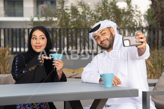 قضاء أوقات ممتعة، زوجان عربيان خليجيان اماراتيان مبتسمان يجلسان في المقهى ، يستمتعان بشرب القهوة الساخنة ، رجل عربي يقوم بإلتقاط صورة سيلفي تذكارية له و لزوجته بسعادة ، قضاء شهر العسل