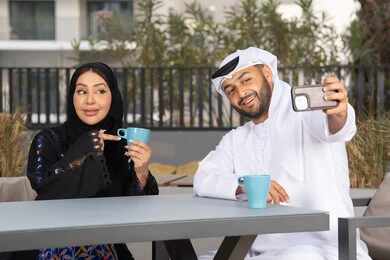 قضاء أوقات ممتعة، زوجان عربيان خليجيان اماراتيان مبتسمان يجلسان في المقهى ، يستمتعان بشرب القهوة الساخنة ، رجل عربي يقوم بإلتقاط صورة سيلفي تذكارية له و لزوجته بسعادة ، قضاء شهر العسل