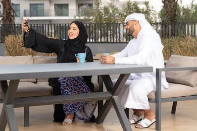 زوجان عربيان خليجيان اماراتيان مبتسمان يجلسان في المقهى ، يستمتعان بشرب القهوة الساخنة ، سيدة عربية تقوم بإلتقاط صورة سيلفي تذكارية لها و لزوجها بسعادة ، صورة للذكرى ، قضاء شهر العسل ، قضاء أوقات ممتعة