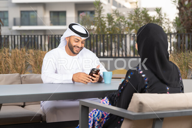 استخدام الهاتف المحمول،  دمج القنيات الحديثة فى الحياة اليومية ،رجل عربي يمسك بيده الجوال ، ايماءات السرور والسعادة ، الاستمتاع بشرب المشروبات الساخنه ، مفهوم المودة والالفة ، قضاء أوقات ممتعة، زوجان عربيان خليجيان اماراتيان مبتسمان يجلسان في المقهى