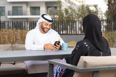 Using a mobile phone, integrating modern technology into daily life, an Arab man holding a mobile phone, gestures of pleasure and happiness, enjoying a hot drink, the concept of affection and intimacy, having a good time, a smiling Emirati Gulf Arab couple sitting in a cafe