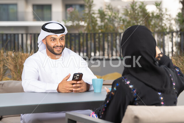 دمج القنيات الحديثة فى الحياة اليومية ،رجل عربي يستخدم الجوال ، ايماءات السرور والسعادة ، الاستمتاع بشرب المشروبات الساخنه ، مفهوم المودة والالفة ، قضاء أوقات ممتعة، زوجان عربيان خليجيان اماراتيان مبتسمان يجلسان في المقهى