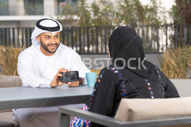 Gestures of pleasure and happiness, honeymoon, enjoying hot drinks, Arab man showing his wife a souvenir photo, concept of affection and intimacy, spending a pleasant time, smiling Arab Gulf Emirati couple sitting in a cafe