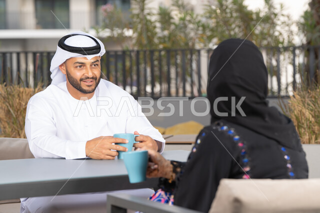 قضاء أوقات ممتعة ورومانسية، ايماءات السرور والسعادة ، الاستمتاع بشرب المشروبات الساخنه ، مفهوم المودة والالفة ، زوجان عربيان خليجيان اماراتيان مبتسمان يجلسان في المقهى