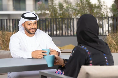 قضاء أوقات ممتعة ورومانسية، ايماءات السرور والسعادة ، الاستمتاع بشرب المشروبات الساخنه ، مفهوم المودة والالفة ، زوجان عربيان خليجيان اماراتيان مبتسمان يجلسان في المقهى