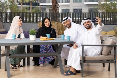 A happy family atmosphere full of love, the concept of intimacy and love between family members, an Emirati man taking commemorative selfies via mobile phone, family entertainment activities and events, gestures of pleasure and happiness, entertainment and recreation during holidays, enjoying drinking hot drinks, an Arab Gulf Emirati family sitting in a cafe