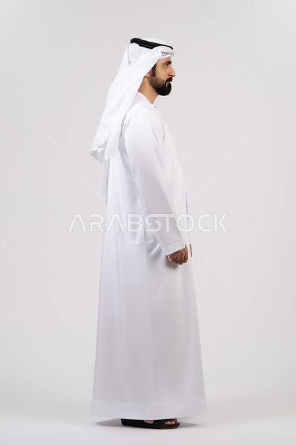 The concept of masculinity and strength, elegance and interest in outward appearance, a portrait of an Emirati Gulf Arab man wearing the Emirati kandura and ghutra, looking at something, standing straight and confident, full body length, white background