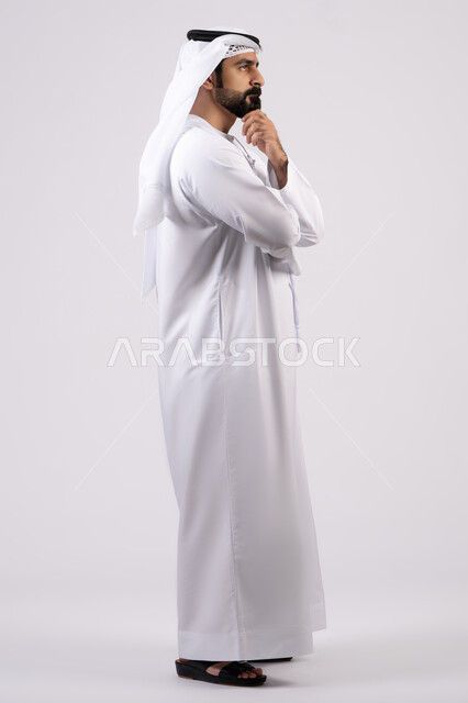 Thinking and trying to find a solution to problems, looking carefully and planning for the future, absent-mindedness and making decisions with focus, portrait of a young Emirati Gulf Arab man wearing a ghutra and kandura looking at something, white background