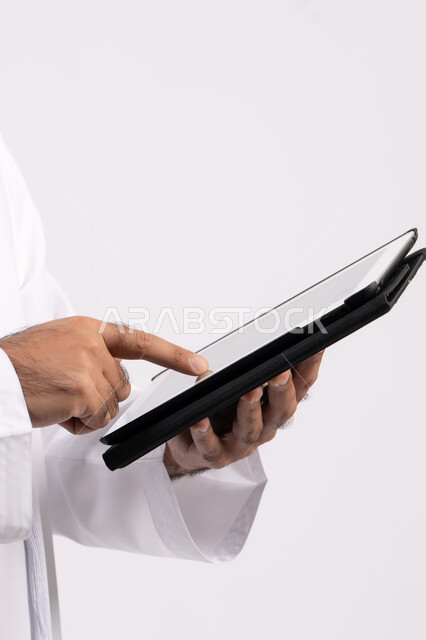 استخدام التكنولوجيا الحديثة ،بورتريه ليد رجل عربي خليجي اماراتية ترتدي الكندورة والغترة يقوم باستخدام الجهاز اللوحي ،ايماءات التفاعل والاندماج , دمج التقنيات الحديثة في الحياة العملية، عرض شاشة سوداء فارغة، صورة للجسم بالكامل، خلفية بيضاء
