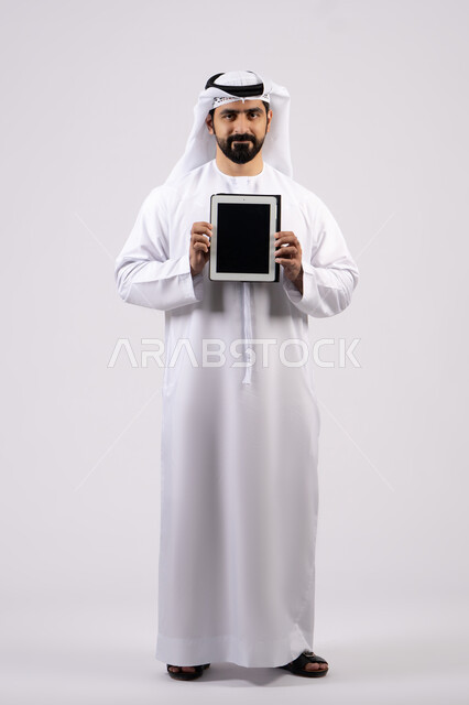 Empty black screen, gestures of interaction and integration, integration of modern technologies in practical life, portrait of an Emirati Gulf Arab man wearing a kandura and ghutra using a tablet, full body image, white background