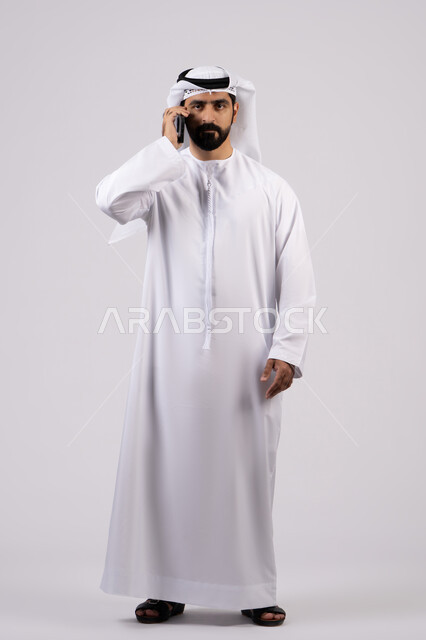 Making phone calls, pointing at mobile phone screen, chatting with family and friends, integrating modern technology into daily life, making a video call, portrait of an Emirati Gulf Arab man wearing a kandura holding a mobile phone, white background.
