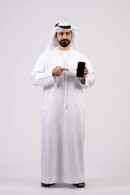 الاشارة الى شاشة الهاتف المحمول ، الدردشة مع الاهل والاصدقاء ، دمج التقنيات والتكنولوجيا الحديثة بالحياة اليومية،  اجراء مكالمة فيديو ، بورتريه لرجل عربي خليجي اماراتي يرتدي الكندورة يمسك الجوال ، خلفية بيضاء.