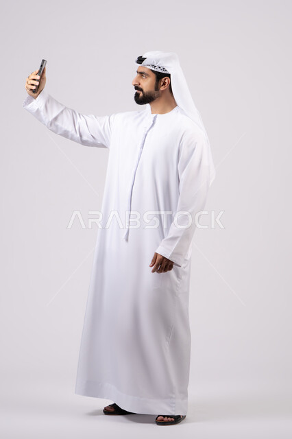 دمج التقنيات والتكنولوجيا الحديثة بالحياة اليومية،  التواصل مع الاهل والاصدقاء، اجراء مكالمة فيديو ،التقاط صور السيلفي باستخدام الهاتف المحمول، توثيق اللقطات الشخصية التذكارية، بورتريه لرجل عربي خليجي اماراتي يرتدي الكندورة يمسك الجوال ، خلفية بيضاء.