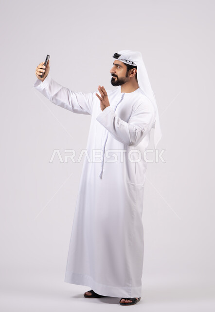 التواصل مع الاهل والاصدقاء، اجراء مكالمة فيديو ،التقاط صور السيلفي باستخدام الهاتف المحمول، دمج التقنيات والتكنولوجيا الحديثة بالحياة اليومية، توثيق اللقطات الشخصية التذكارية، بورتريه لرجل عربي خليجي اماراتي يرتدي الكندورة يمسك الجوال ، خلفية بيضاء.