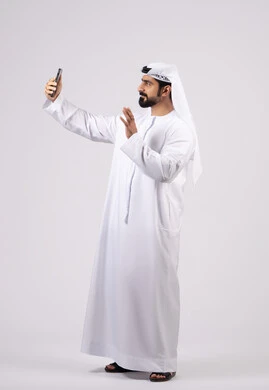 Communicating with family and friends, making a video call, taking selfies with a mobile phone, integrating modern technology into daily life, documenting personal memories, a portrait of an Emirati Gulf Arab man wearing a kandura holding a mobile phone, white background.