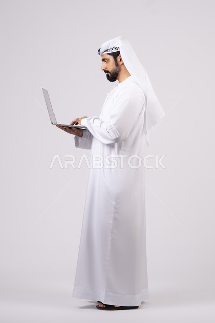 تصفح مواقع التواصل الإجتماعي ،ادارة شؤون الاعمال وتنظيمها، استخدام الأجهزة التقنية الحديثة والمتطورة، العمل عن بعد،  بورتريه لرجل عربي خليجي اماراتي يرتدي الكندورة والغترة البيضاء يستخدم الحاسوب المحمول، اجراء مكالمات الفيديو، خلفية بيضاء