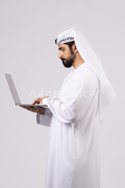 Working remotely, browsing social media, managing and organizing business affairs, using modern and advanced technical devices, portrait of an Emirati Gulf Arab man wearing a white kandura and ghutra using a laptop, making video calls, white background