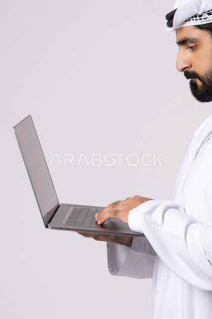ادارة شؤون الاعمال وتنظيمها، العمل عن بعد، تصفح مواقع التواصل الإجتماعي ،استخدام الأجهزة التقنية الحديثة والمتطورة، بورتريه لرجل عربي خليجي اماراتي يرتدي الكندورة والغترة البيضاء يستخدم الحاسوب المحمول، اجراء مكالمات الفيديو، خلفية بيضاء