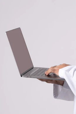 اجراء بث مباشر، ادارة شؤون الاعمال وتنظيمها، العمل عن بعد، تصفح مواقع التواصل الإجتماعي ،استخدام الأجهزة التقنية الحديثة والمتطورة، بورتريه مقرب ليد رجل عربي خليجي اماراتي يرتدي الكندورة والغترة البيضاء يستخدم الحاسوب المحمول، اجراء مكالمات الفيديو، خلفية بيضاء