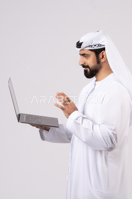 ادارة شؤون الاعمال وتنظيمها، استخدام الأجهزة التقنية الحديثة والمتطورة، العمل عن بعد،  تصفح مواقع التواصل الإجتماعي ، بورتريه لرجل عربي خليجي اماراتي يرتدي الكندورة والغترة البيضاء يستخدم الحاسوب المحمول، اجراء مكالمات الفيديو، خلفية بيضاء