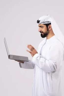 ادارة شؤون الاعمال وتنظيمها، استخدام الأجهزة التقنية الحديثة والمتطورة، العمل عن بعد،  تصفح مواقع التواصل الإجتماعي ، بورتريه لرجل عربي خليجي اماراتي يرتدي الكندورة والغترة البيضاء يستخدم الحاسوب المحمول، اجراء مكالمات الفيديو، خلفية بيضاء