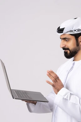Using modern and advanced technical devices, working remotely, browsing social networking sites, portrait of an Emirati Gulf Arab man wearing a white kandura and ghutra using a laptop, making video calls, managing and organizing business affairs, white background