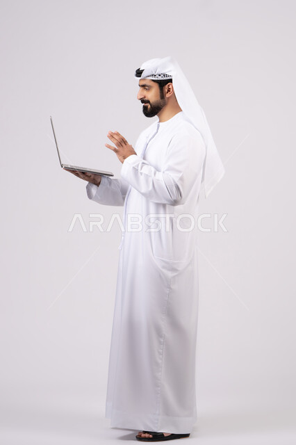 ادارة شؤون الاعمال وتنظيمها، استخدام الأجهزة التقنية الحديثة والمتطورة،العمل عن بعد،  تصفح مواقع التواصل الإجتماعي ، بورتريه لرجل عربي خليجي اماراتي يرتدي الكندورة والغترة البيضاء يستخدم الحاسوب المحمول، خلفية بيضاء