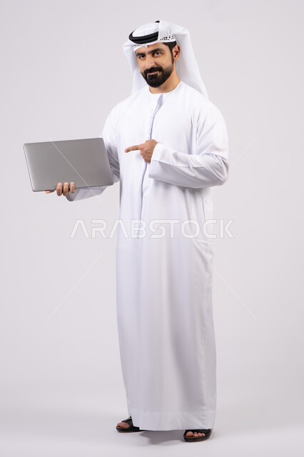 الاشارة الى جهاز اللابتوب، ادارة شؤون الاعمال وتنظيمها، العمل عن بعد، تصفح مواقع التواصل الإجتماعي ،استخدام الأجهزة التقنية الحديثة والمتطورة، بورتريه  لرجل عربي خليجي اماراتي يرتدي الكندورة والغترة البيضاء يستخدم الحاسوب المحمول، اجراء مكالمات الفيديو، خلفية بيضاء