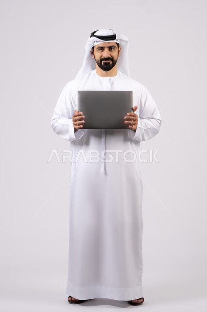العمل عن بعد، ادارة شؤون الاعمال وتنظيمها، استخدام الأجهزة التقنية الحديثة والمتطورة، تصفح مواقع التواصل الإجتماعي ، بورتريه لرجل عربي خليجي اماراتي يرتدي الكندورة والغترة البيضاء يستخدم الحاسوب المحمول، خلفية بيضاء