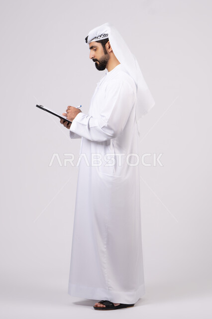 تسجيل الملاحظات والمعلومات على الورق,بورتريه لشاب عربي خليجي اماراتي يرتدي الكندورة والغترة يمسك مجموعة من الاوراق البيضاء وقلم, كتابة المهام والأهداف اليومية، تنظيم وإدارة الأعمال ،تدوين المهام اليومية ، خلفية بيضاء