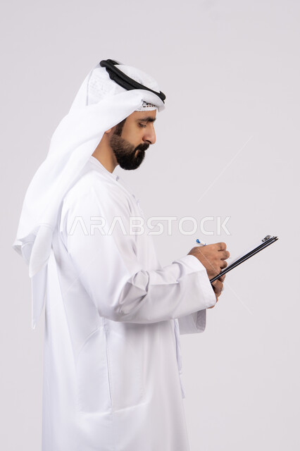 تنظيم وإدارة الأعمال ، تدوين وتسجيل الملاحظات والمعلومات على الورق, بورتريه لشاب عربي خليجي اماراتي يرتدي الكندورة والغترة يقوم بكتابة المهام والأهداف اليومية, خلفية بيضاء