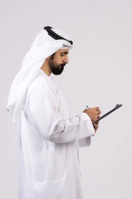 تنظيم وإدارة الأعمال ، تدوين وتسجيل الملاحظات والمعلومات على الورق, بورتريه لشاب عربي خليجي اماراتي يرتدي الكندورة والغترة يقوم بكتابة المهام والأهداف اليومية, خلفية بيضاء
