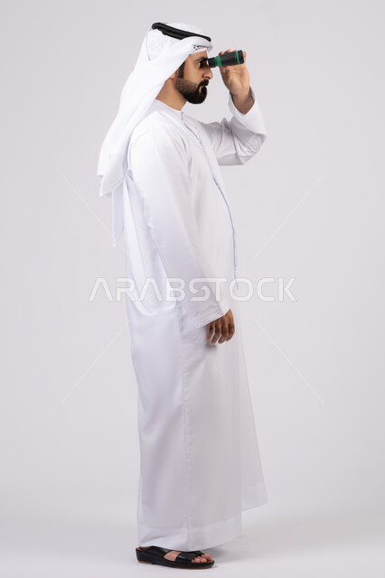حب المغامرة والاستكشاف، رؤية الاشياء البعيدة عبر التلسكوب، بورتريه لرجل عربي خليجي إماراتي يرتدي الكندورة والغترة ينظر من خلال المنظار ثنائي العينين، ايماءات وتعابير الدهشة والمفاجئة، خلفية بيضاء