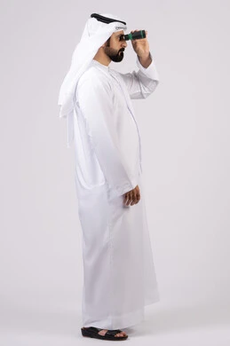 حب المغامرة والاستكشاف، رؤية الاشياء البعيدة عبر التلسكوب، بورتريه لرجل عربي خليجي إماراتي يرتدي الكندورة والغترة ينظر من خلال المنظار ثنائي العينين، ايماءات وتعابير الدهشة والمفاجئة، خلفية بيضاء