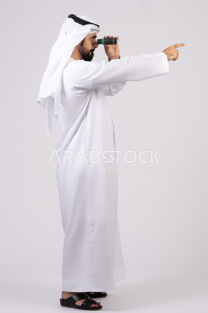 الاشارة الى شئ بعيد، بورتريه لرجل عربي خليجي إماراتي مبتسم يرتدي الكندورة والغترة ينظر من خلال المنظار ثنائي العينين، حب المغامرة والاستكشاف، رؤية الاشياء البعيدة عبر التلسكوب،ايماءات وتعابير الدهشة والمفاجئة، خلفية بيضاء