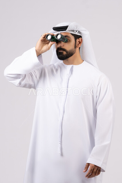 حب المغامرة والاستكشاف، رؤية الاشياء البعيدة عبر التلسكوب،ايماءات وتعابير الدهشة والمفاجئة، بورتريه لرجل عربي خليجي إماراتي مبتسم يرتدي الكندورة والغترة ينظر من خلال المنظار ثنائي العينين، خلفية بيضاء