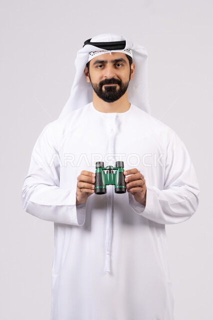 Seeing distant objects through a telescope, gestures and expressions of astonishment and surprise, portrait of a smiling Emirati Gulf Arab man wearing a kandura and ghutra looking through binoculars, love of adventure and exploration, white background