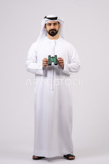 Gestures and expressions of surprise and amazement, seeing distant objects through a telescope, portrait of a smiling Emirati Gulf Arab man wearing a kandura and ghutra looking through binoculars, love of adventure and exploration, white background
