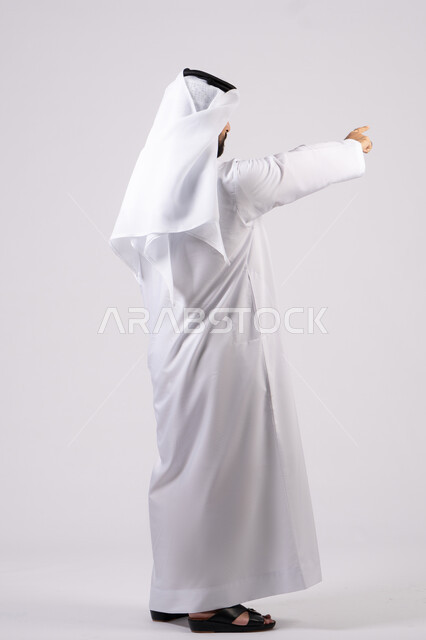 تحديد موقع شيء معين، إشارات وحركات يدين تدل على هذا المكان (هنا)، بورتريه من الخلف لرجل عربي خليجي اماراتي يرتدي الكندورة والغترة البيضاء يشير باصبع السبابة إلى الأعلى، مفهوم الأناقة والاهتمام بالمظهر الخارجي، خلفية بيضاء