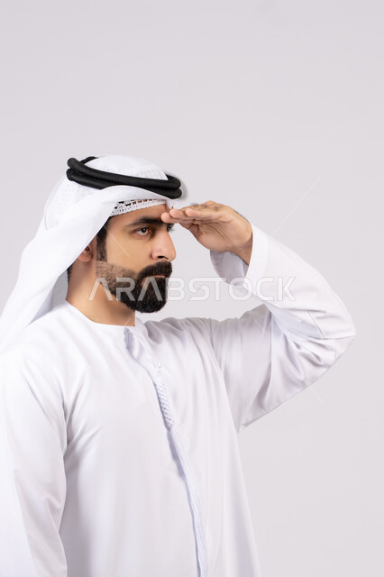 تعابير محاولة رؤية الأشياء البعيدة, مفهوم التطلع للمستقبل, النظر لمكان ما, بورتريه لرجل عربي خليجي إماراتي يرتدي الكندورة الغترة البيضاء يرفع يده إلى رأسه بإيماءات البحث عن شيء محدد, خلفية بيضاء