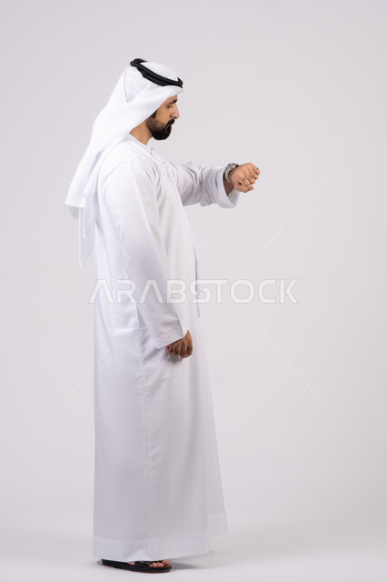 The concept of the importance of time and commitment to appointments, a portrait of an Emirati Gulf Arab man wearing a dakandura and ghutra looking at his personal watch, gestures of the importance of commitment to time and completing tasks and work accurately, white background
