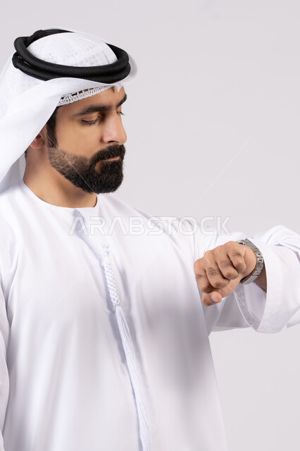 مفهوم اهميه الوقت والالتزام بالمواعيد، بورتريه لرجل عربي خليجي اماراتي يرتدي الدكندورة والغترة ينظر الى ساعته الشخصيه بايماءات اهميه الالتزام بالوقت وإنجاز المهامات والاعمال بدقه، خلفيه بيضاء